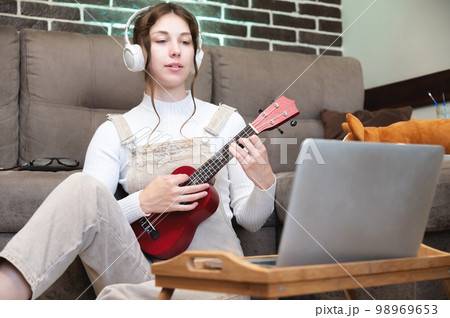 young beautiful woman is sitting on the floor at home and learning to play the guitar or ukulele, standing next to a laptop and a cup of coffee. distance learning concept 98969653