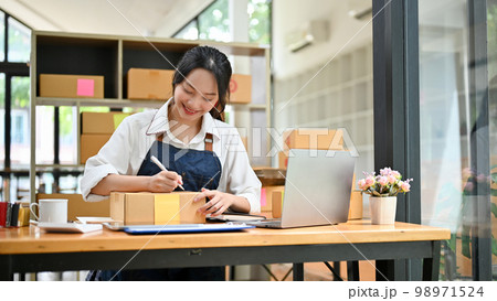 Asian female small online business owner writing customer's address on a cardboard box 98971524