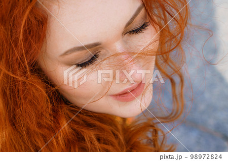 Close-up portrait young caucasian woman with red curly hair in red coat in wind Close-up portrait young caucasian woman with red curly hair in red coat in wind 98972824