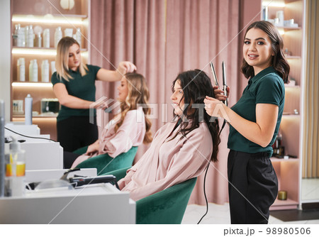 Portirait of professional hairdresser doing hairstyle for woman in beauty salon. Smiling female hairstylist curling client hair with curling iron device while colleague combing woman hair. 98980506