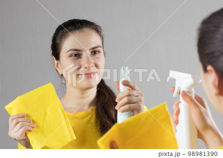 A beautiful woman in a yellow bright T-shirt smiles and cleaning mirror with a water squeegee with a scraper. Means for cleaning windows and glass. Cleaning service. Maid cleaning at home. 98981390