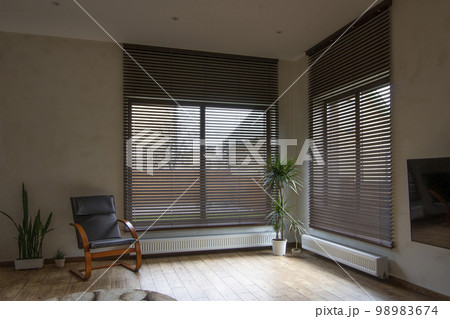 Wooden blinds on large windows in the interior. Living room with armchair and houseplants near windows with wood blinds. Motorized jalousie in the smart house. 98983674