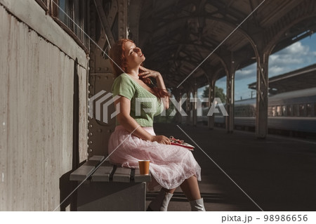 redhead young woman in green bun and pink skirt dreamily sits with glass of coffee at train station 98986656