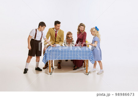 Friendly happy family, parents and kids standing around table isolated on white background. Concept of holidays, traditions, retro style, fashion, elegance, 60s, 70s, family. Friendly happy family, parents and kids standing around table isolated on white background. Concept of holidays, traditions, retro style, fashion, elegance, 60s, 70s, family. 98986673