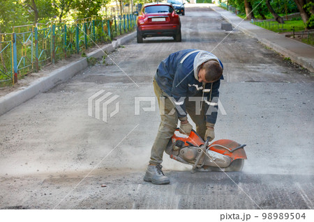 A worker cuts worn-out asphalt with a petrol cutter in a cloud of dust against the backdrop of a road being repaired. 98989504