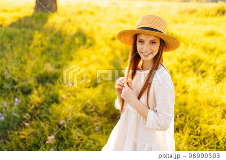 Portrait of beautiful red-haired young woman in straw hat and white dress standing posing on beautiful meadow of green grass looking at camera, on background of bright sunlight at summer day. 98990503