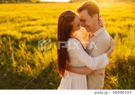 Portrait of cheerful young couple in love standing hugging with closed eyes on beautiful green meadow in summer evening during golden sunset with soft sunlight. Concept of romantic date love outdoors. Portrait of cheerful young couple in love standing hugging with closed eyes on beautiful green meadow in summer evening during golden sunset with soft sunlight. Concept of romantic date love outdoors. 98990504