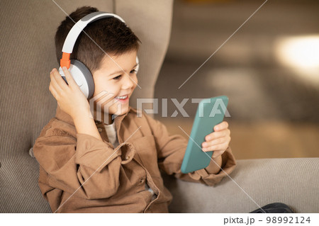 Happy european little boy in casual and wireless headphones watch video on smartphone, sits in armchair Happy european little boy in casual and wireless headphones watch video on smartphone, sits in armchair 98992124