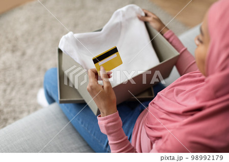 Delivery service and online payment. Black lady in hijab opening box with new t-shirt and holding credit card 98992179