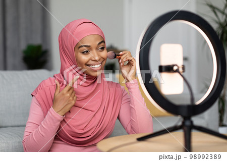 Cheerful black muslim woman recording cosmetics product review, applying powder, filming video for her vlog using phone 98992389