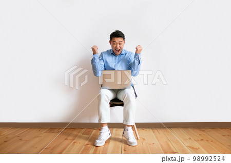 Overjoyed middle aged asian man with laptop computer sitting on chair, celebrating great deal, making YES gesture 98992524