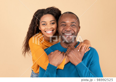 Portrait of happy young black woman hugging her middle aged husband from behind over peach studio background 98992525