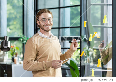 A young designer, programmer, freelancer man is standing in the office, holding a tablet, drawing a progress graph on a transparent board. Glue colored, stickers, hints. He looks at the camera, smiles A young designer, programmer, freelancer man is standing in the office, holding a tablet, drawing a progress graph on a transparent board. Glue colored, stickers, hints. He looks at the camera, smiles 98994174