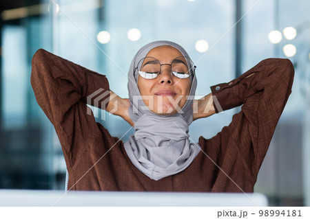 Portrait of a young businesswoman in hijab and glasses sitting in the office at the table in front of the computer. She put her hands behind head, closed her eyes, resting, taking a break, smiling. 98994181