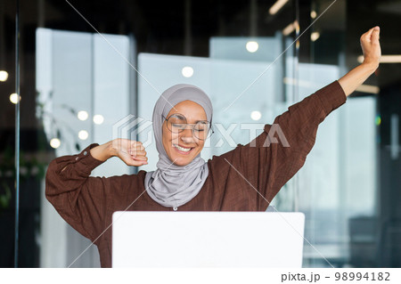 Portrait of young Arab businesswoman in hijab and glasses sitting in office at table with laptop. She is tired, has finished her work day, has done her work, stretches her hands up, smiles, rests. 98994182