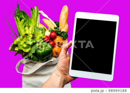 Online shopping mobile app holding by a hand with blurred background of fresh vegetables and healthy food on isolated background Online shopping mobile app holding by a hand with blurred background of fresh vegetables and healthy food on isolated background 98994188