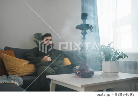Bearded man is smoking hookah at home and blowing cloud of smoke, copy space and empty space for text - chill time and resting concept 98994695
