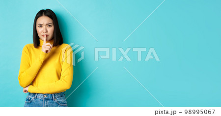 Disappointed asian woman telling to be quiet, scolding loud person with hush gesture, shushing at camera and frowning upset, standing over blue background 98995067