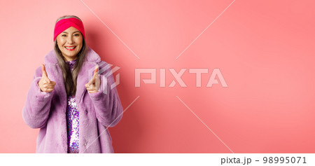 Fashion and shopping concept. Cool asian senior woman in stylish fake fur coat pointing fingers at camera, asking you to check out promo offer, standing over pink background Fashion and shopping concept. Cool asian senior woman in stylish fake fur coat pointing fingers at camera, asking you to check out promo offer, standing over pink background 98995071