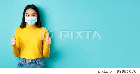 Covid-19, social distancing and pandemic concept. Supportive asian woman recommend promotion, wearing medical mask and showing thumbs up, standing over blue background Covid-19, social distancing and pandemic concept. Supportive asian woman recommend promotion, wearing medical mask and showing thumbs up, standing over blue background 98995076