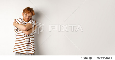Reluctant young man with messy red hair unwilling do something, hugging himself and grimacing, standing over white background 98995084