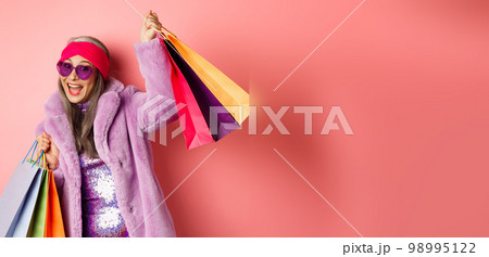 Funky and cool asian senior woman in fashionable clothes dancing while going shopping on sales, holding shop paper bags and having fun, pink background 98995122