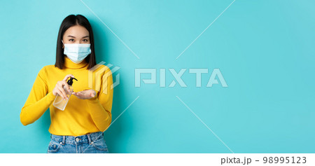 Covid-19, social distancing and pandemic concept. Smiling asian woman in medical mask using preventive measures from coronavirus, using hand sanitizer, standing over blue background 98995123