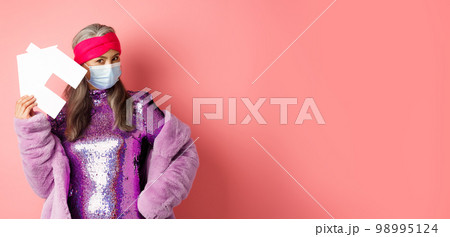 Covid-19 and real estate concept. Cool and stylish asian elderly woman in disco dress and medical mask showing paper house, standing over pink background 98995124