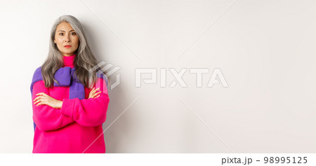 Serious and strict asian grandmother looking angry at camera, cross arms on chest, standing over white background disappointed 98995125
