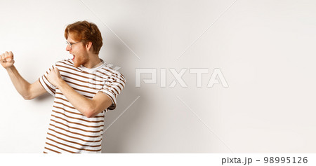 Profile of funny young man with red hair and beard, raising fists for fight, shadow boxing and shouting, standing over white background 98995126