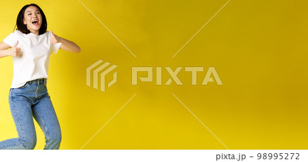 Full size of happy young asian woman jumping from joy, showing thumbs up in approval, posing over yellow background in jeans and casual white t-shirt 98995272