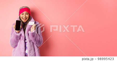Online shopping and fashion concept. Beautiful asian mature woman showing plastic credit card and blank phone screen, smiling happy, pink background Online shopping and fashion concept. Beautiful asian mature woman showing plastic credit card and blank phone screen, smiling happy, pink background 98995423