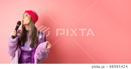 Gorgeous asian senior woman singing karaoke in mic, perform song and looking happy, standing over pink background Gorgeous asian senior woman singing karaoke in mic, perform song and looking happy, standing over pink background 98995424