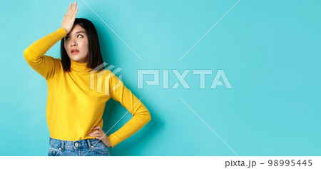 Emotions and lifestyle concept. Annoyed asian girl roll eyes and face palm, standing bothered in yellow pullover against blue background 98995445