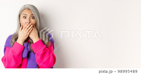 Close up of shocked senior lady gasping and covering mouth, staring at camera speechless, witness something, standing over white background 98995489