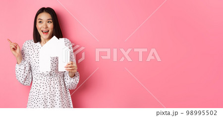Real estate. Excited asian woman showing paper house model, pointing and looking left at copy space, standing over pink background Real estate. Excited asian woman showing paper house model, pointing and looking left at copy space, standing over pink background 98995502