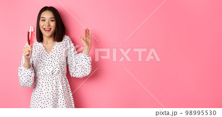 Celebration. Beautiful asian woman drinking champagne and smiling, standing in dress over pink background 98995530
