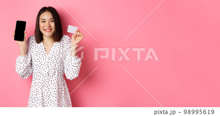 Joyful korean woman showing smartphone screen and credit card, paying for internet order, demonstrating online shopping app, standing over pink background Joyful korean woman showing smartphone screen and credit card, paying for internet order, demonstrating online shopping app, standing over pink background 98995619