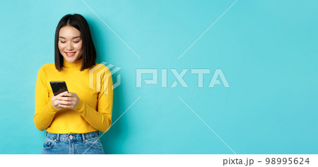 Attractive asian woman reading smartphone screen and smiling, social networking with mobile phone, standing over blue background 98995624