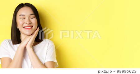 Close up of romantic asian woman dreaming of something cute, close eyes and hold hands near face, imaging beautiful thing, standing over yellow background 98995625