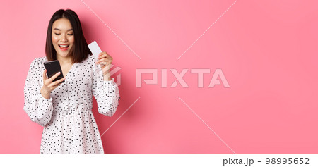Attractive young asian woman order online, holding credit card and mobile phone, making internet purchase, standing happy over pink background 98995652