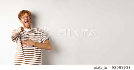 Young man with ginger hair and beard pointing finger at camera and laughing, making fun of someone hilarious, standing over white background 98995653
