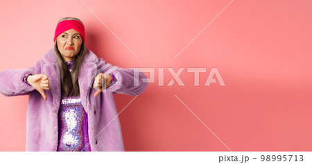Fashion and shopping concept. Disappointed stylish senior woman showing thumbs-down, looking displeased and judgemental, being dissatisfied, standing over pink background Fashion and shopping concept. Disappointed stylish senior woman showing thumbs-down, looking displeased and judgemental, being dissatisfied, standing over pink background 98995713