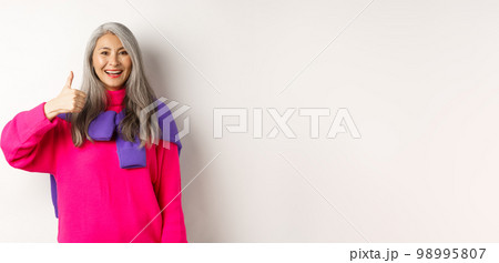 Portrait of happy and satisfied asian senior woman showing thumb-up, approval and like something, smiling and praising product, standing over white background Portrait of happy and satisfied asian senior woman showing thumb-up, approval and like something, smiling and praising product, standing over white background 98995807