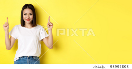 Beauty and fashion concept. Beautiful asian woman in white t-shirt pointing fingers up, standing over yellow background Beauty and fashion concept. Beautiful asian woman in white t-shirt pointing fingers up, standing over yellow background 98995808
