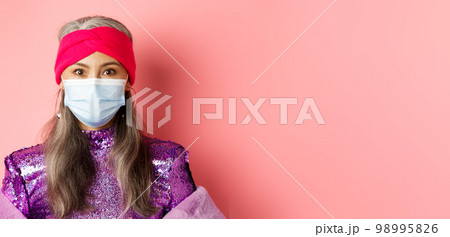 Covid-19, virus and social distancing concept. Close-up of stylish asian senior woman in medical mask and shiny dress looking at camera, standing over pink background 98995826