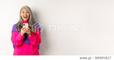Surprised asian woman smiling at camera after reading promotion on smartphone, standing with mobile phone over white background 98995827