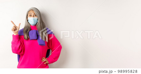 Covid-19, quarantine and lockdown concept. Amazed asian lady in face mask looking at camera impressed, showing logo on upper left corner, pointing at banner, white background 98995828