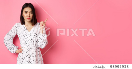 Sad and gloomy asian girl frowning, pointing finger at upper right corner and staring at camera unhappy, standing over pink background 98995938