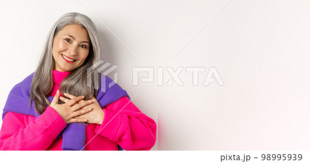 Close up of stylish asian mother holding hands on heart, looking touched and heartfelth, saying thank you, standing over white background 98995939